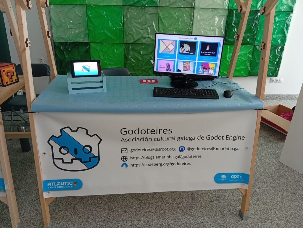 Foto do posto, cun ordenador en marcha con xogos feitos en Godot. O posto ten unha pancarta co nome da asociación, email, dirección do blog e redes.