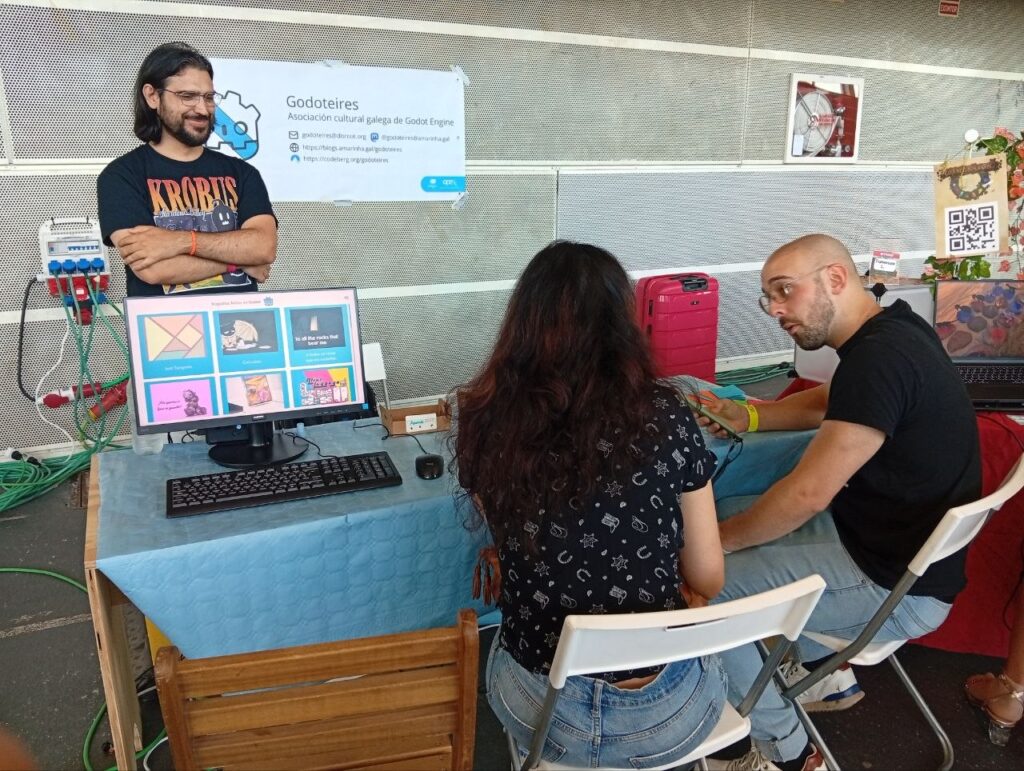 Persoas xogando no stand da aso. O noso compi observa a ambos detrás do stand, no que tamén hai unha pancarta co logo e as redes da asociación