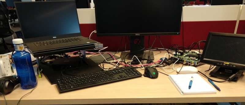 Dúas pantallas e un ordenador portátil, un teclado e ratón, unha placa electrónica, un caderno e dous bolígrafos, unha botellas e outros pequenos obxectos. Todo sobre unha mesa de madeira e contra unha parede branca e vermella.
