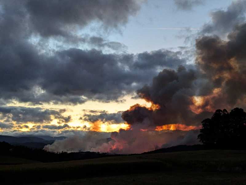 Un incendio. O ceo con nubes pero bastante despexado vese cuberto na parte dereita por unha fumareda contra a cal se reflexa a luz vermella do lume. Máis escuras vense a ssombras das montañas e das árbores que están máis preto da cámara
