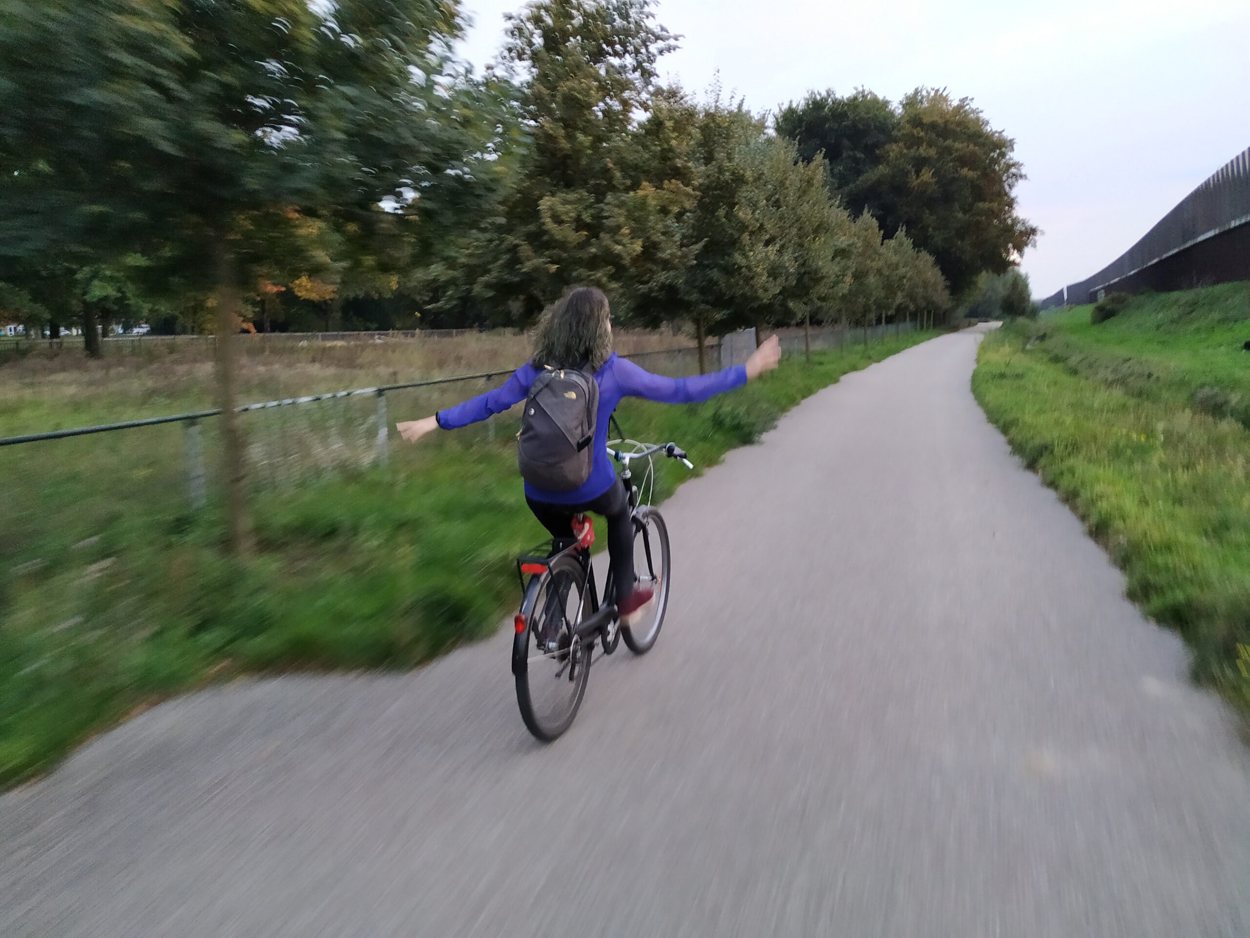 Unha rapaza de espaldas montada en bicicleta holandesa e peladeando sen mans. Atravesa un parque sobre un camiño asfaltado.