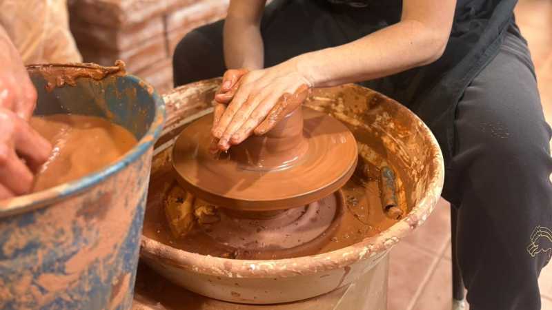 No centro un torno de orfebre sobre o cal traballa o barro unha persoa, só medio corpo se ve con roupa negra. Á esquerda da foto, vese un caldeiro de auga con barro e as mans de outra persoa a remollo.