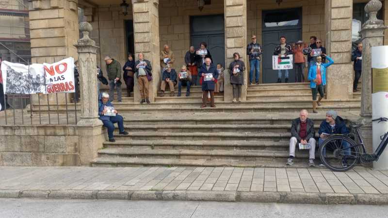Grupo de persoas manifestándose nas escaleiras e soportais do concello de Ribadeo, un edificio de pedra ocre coas portas azuis. Hai pancartas de non a guerra e bandeiras de palestina.